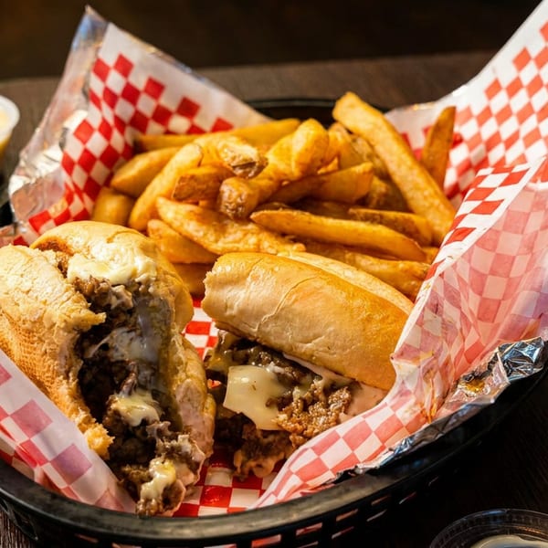Two cheesesteak sandwiches and a pile of french fries in a basket with red and white checkered paper.
