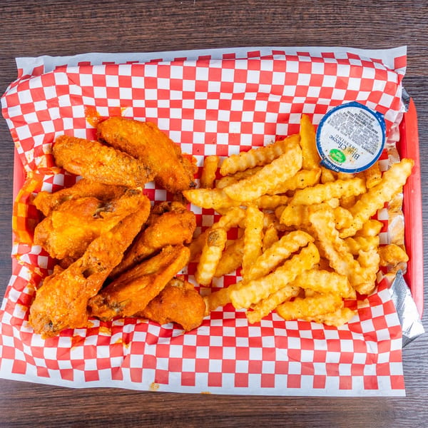 Basket of crispy chicken wings and crinkle-cut fries with dipping sauce on red-and-white checkered paper.