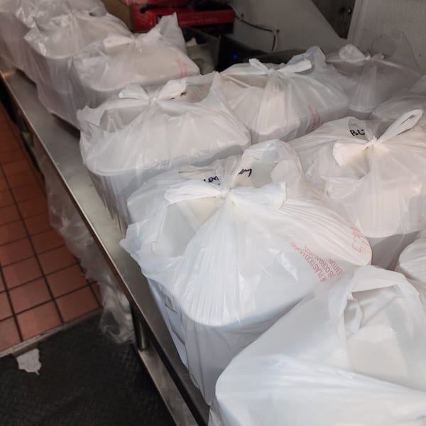 Numerous white takeout containers in plastic bags line a stainless steel counter in a commercial kitchen.