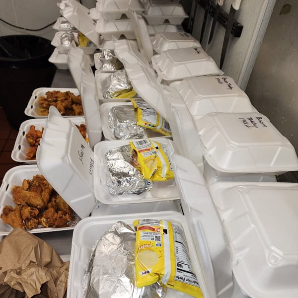 Numerous styrofoam takeout containers filled with chicken wings, foil-wrapped items, and chip bags on a counter.