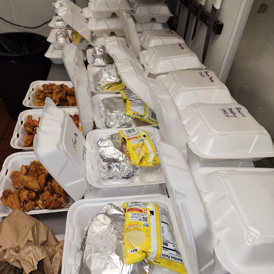 Numerous styrofoam takeout containers filled with chicken wings, foil-wrapped items, and chip bags on a counter.