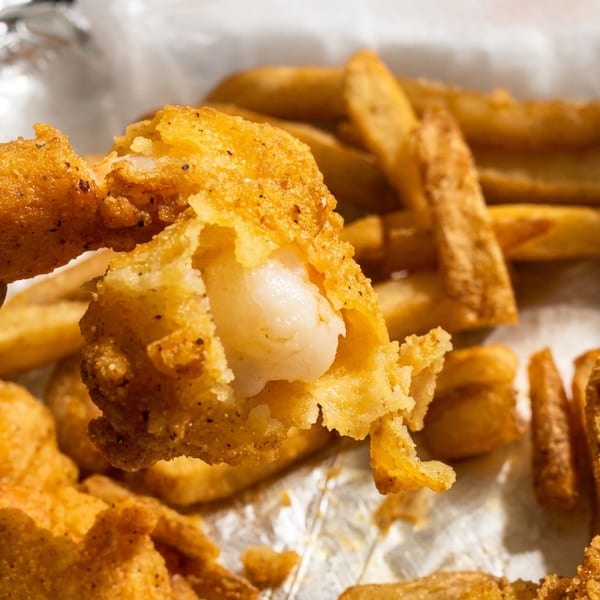 Hand holding a piece of fried shrimp, revealing the white meat inside, with fries in the background.