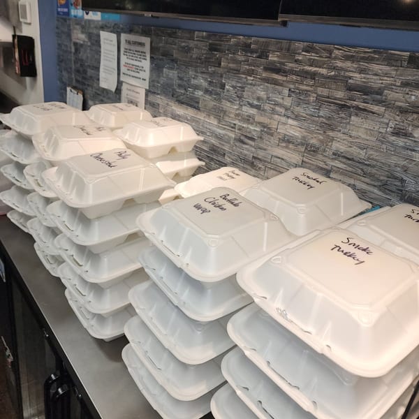 Stacks of white takeout containers with handwritten labels like "Philly Cheesesteak" and "Smoked Turkey" on a counter.