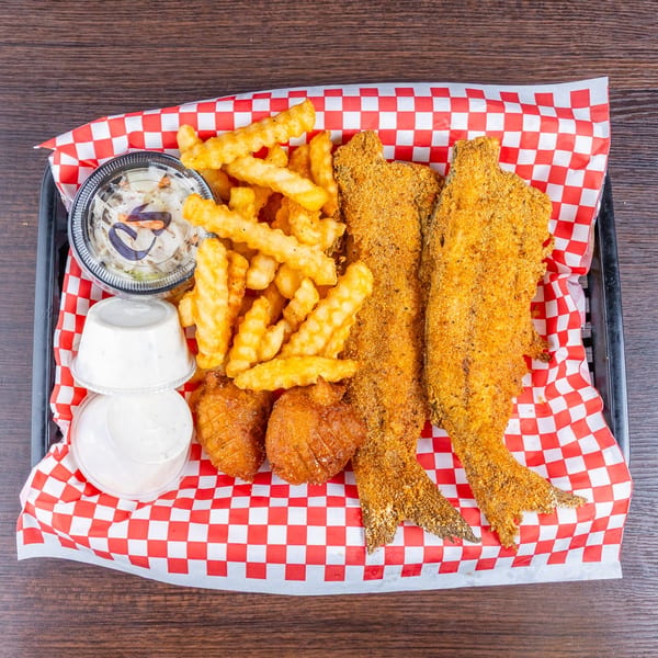 Basket of fried fish, crinkle-cut fries, hush puppies, coleslaw, and dipping sauces on red-and-white checkered paper.