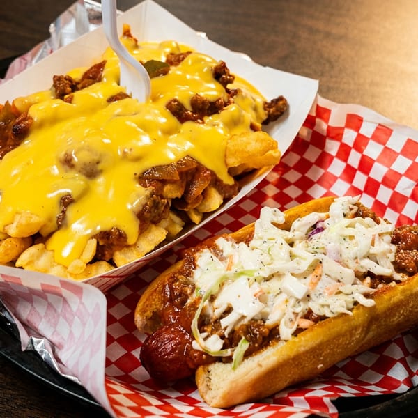 Chili cheese fries and a hot dog with chili and coleslaw on red and white checkered paper.