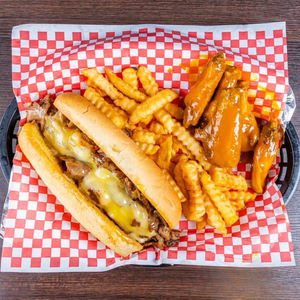 Cheesesteak sandwich with melted cheese, crinkle-cut fries, and saucy chicken wings on red-and-white checkered paper.