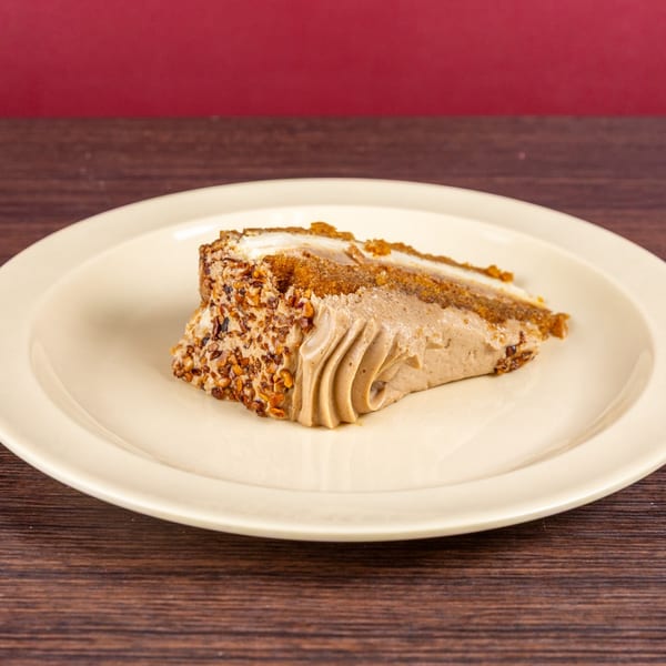 Slice of layered cake with caramel-colored frosting and nut-coated sides on a cream plate atop a wooden table.