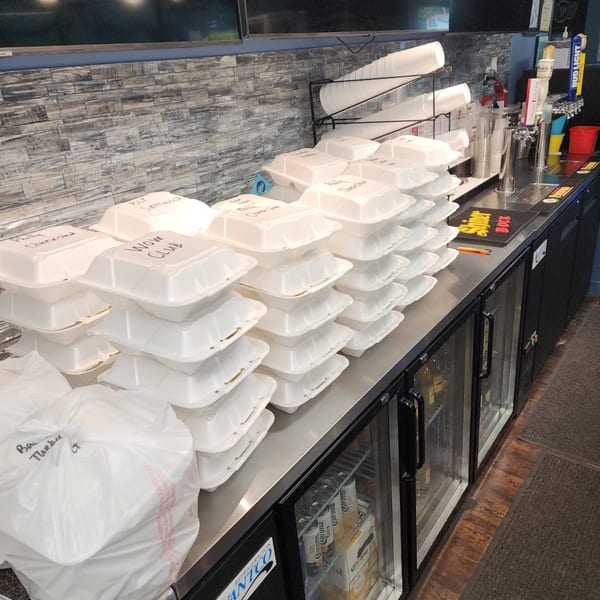 Numerous stacked takeout containers with handwritten labels like "WOW CLUB" and "Philly Cheesesteak" on a counter.