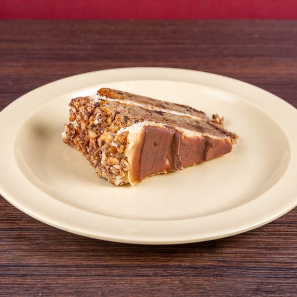 Slice of layered cake with cream filling and nut topping on a cream-colored plate atop a wooden table.